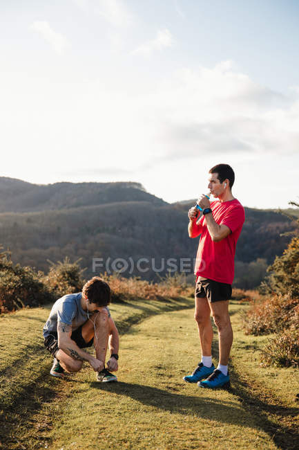 Adult athletic man standing on green trail and drinking water while friend tying running shoes during training in mountains in sunny day — Stock Photo
