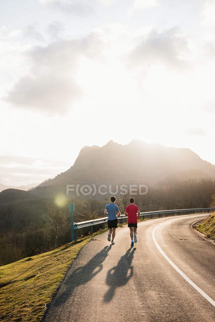 Back view of anonymous active healthy male joggers running together on curved asphalt road with sunlight from behind mountain in background — Stock Photo