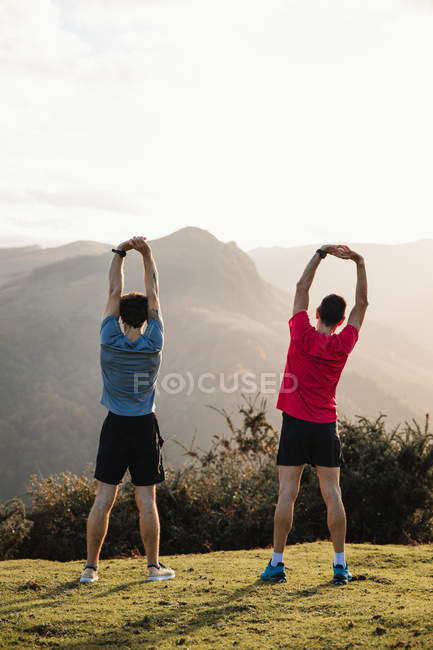 Вид ззаду втомлених чоловічих бігунів у синьо-червоних сорочках, що тягнуться після бігу та тренувань, стоячи на зеленому пагорбі — стокове фото