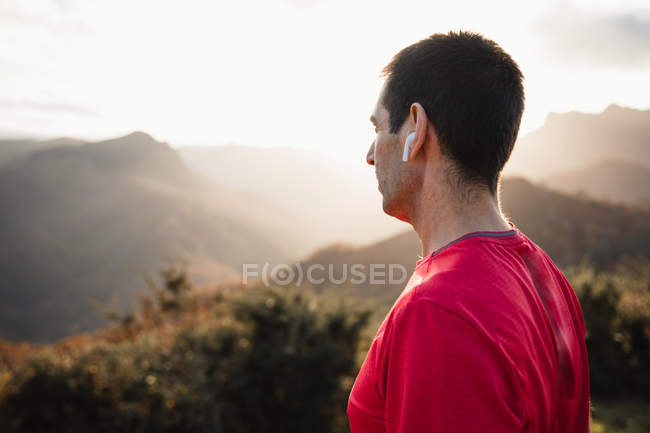 Side view of sportive man in blue and red shirts standing on top of green hill and enjoying landscape while relaxing after running — Stock Photo