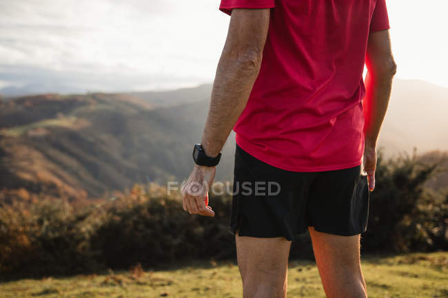 Vue arrière de l'homme sportif en chemises bleues et rouges debout au sommet de la colline verte et profitant du paysage — Photo de stock