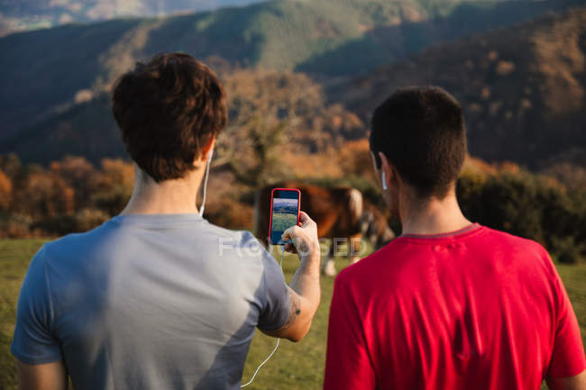 Visão traseira de machos esportivos em pé no topo da colina verde tirando foto com telefone celular de uma vaca no pasto — Fotografia de Stock