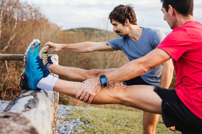 Seitenansicht von müden männlichen Joggern in blauen und roten Hemden, die sich nach dem Laufen und hartem Training auf dem grünen Hügel über den Holzzaun strecken — Stockfoto