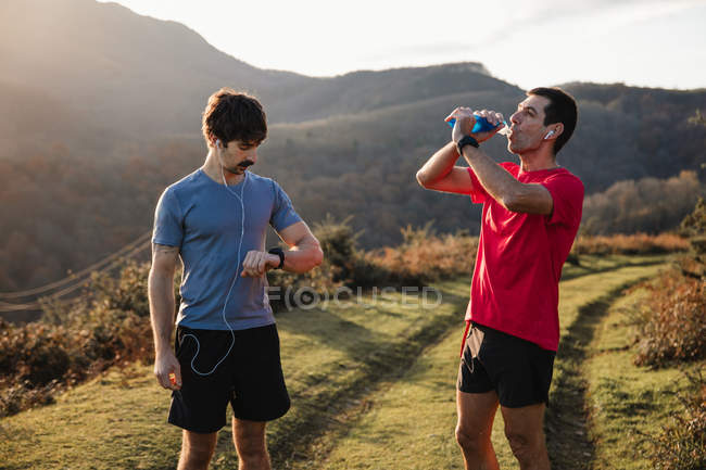 Adult athletic man standing on green trail and drinking water while friend look at the watch during training in mountains in sunny day — Stock Photo