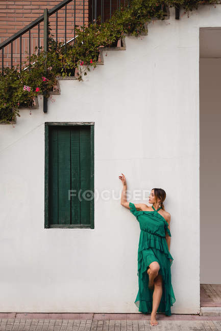 Женщина в зеленом платье опирается на белую стену дома с лестницей и цветами в горшках на улице города — стоковое фото