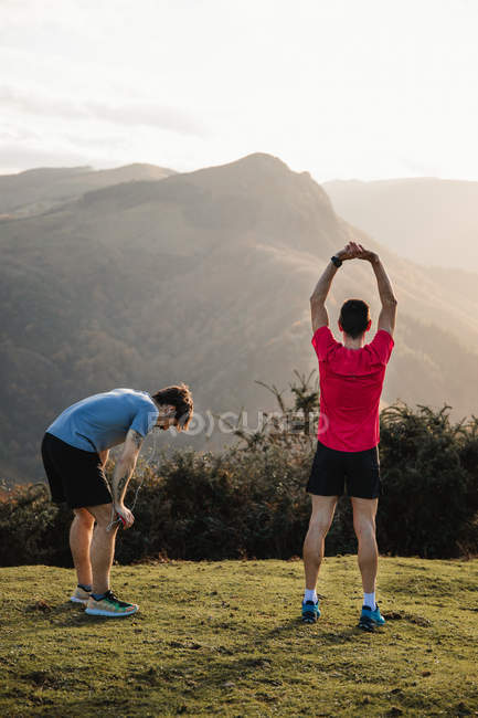 Back view of tired male joggers in blue and red shirts stretching after running and training hard while standing on top of green hill — Stock Photo