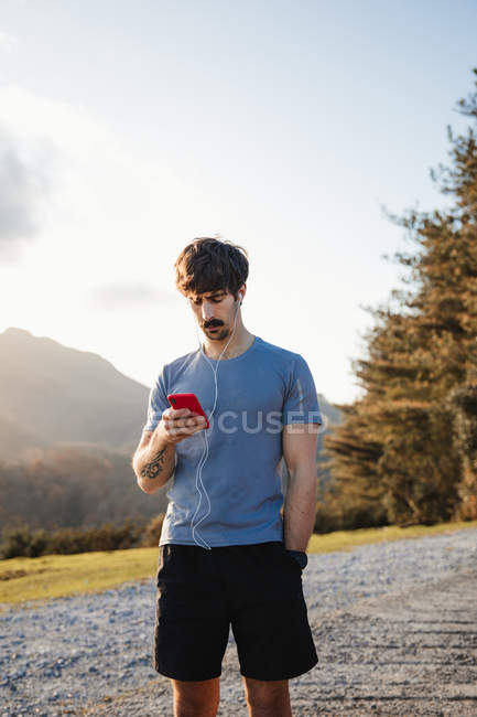 Sportive man standing on asphalt narrow path enjoying mountain landscape while listening to music on mobile phone on earphones — Stock Photo