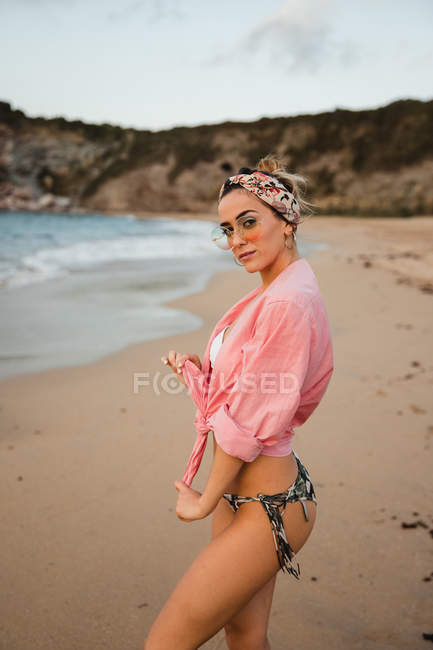 Side view of trendy woman in swimwear and sunglasses tying shirt and looking at camera while standing on sandy beach — Stock Photo