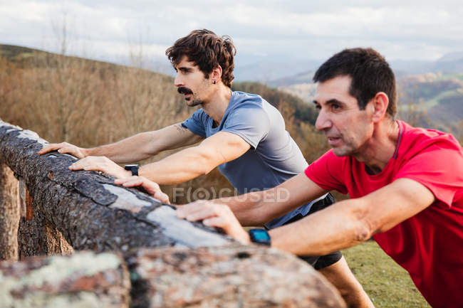 Vista lateral de los joggers masculinos cansados en camisas azules y rojas que se estiran en la cerca de madera después de correr y entrenar duro en la colina verde - foto de stock