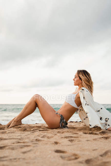 Вид сбоку женщины в бикини и блузке, откидывающейся назад и отводящей взгляд, сидя на песчаном берегу на фоне пасмурного неба — стоковое фото