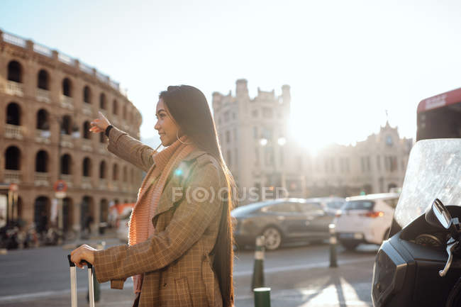 Side view of Asian woman with suitcase smiling and stretching out arm while hailing cab on street of tourist city — Stock Photo