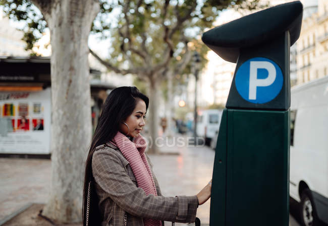 Вид сбоку на азиатскую женщину в стильной одежде с использованием современного паркомата во время поездки по городу — стоковое фото