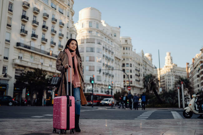 Asian woman with suitcase standing on pavement near road and looking away while visiting city on sunny day — Stock Photo