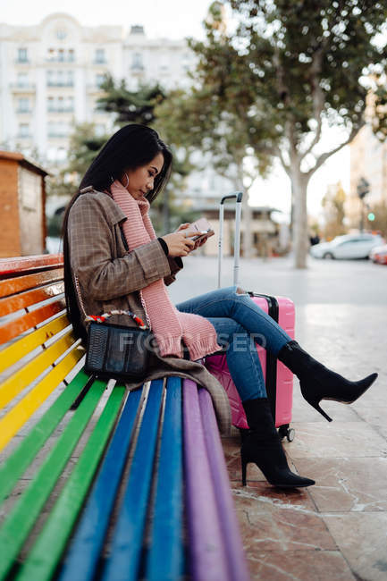 Seitenansicht asiatische Frau mit Koffer sitzt auf Regenbogenbank und surft Smartphone auf der Stadtstraße — Stockfoto
