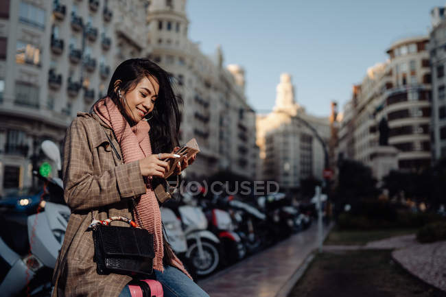 Longitud completa feliz mujer asiática con teléfono inteligente sonriendo y mirando hacia otro lado mientras está sentado en la maleta en la acera en la calle de la ciudad - foto de stock