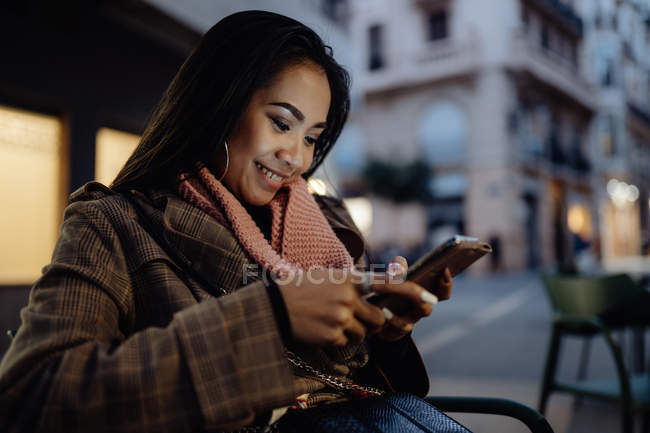Asiatin lächelt und surft auf ihrem Smartphone in den sozialen Medien, während sie sich abends in einem Straßenrestaurant ausruht — Stockfoto