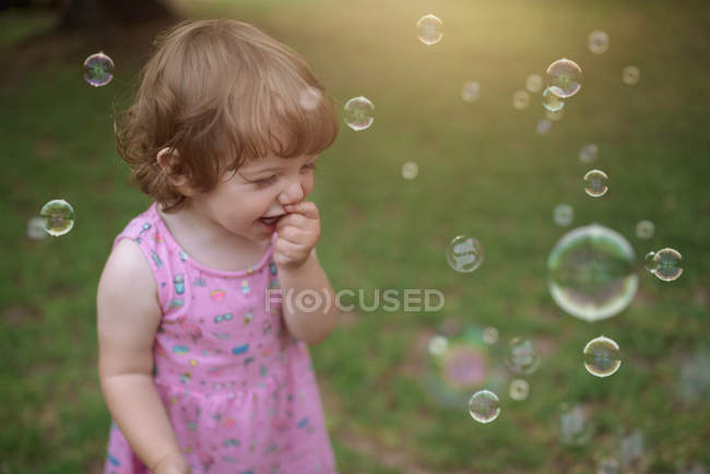 Сверху очаровательный ребенок в розовом платье смеется и захватывает радужные мыльные пузыри на зеленом лугу в парке — стоковое фото