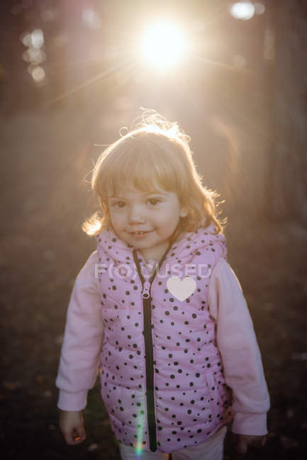 Affascinante bambino allegro in caldo gilet rosa con cuore guardando la fotocamera alla luce del sole nel parco — Foto stock