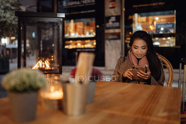 Mujer asiática sonriendo y navegando teléfono inteligente mientras está sentado en la mesa del restaurante cerca de la maleta en la noche en la calle de la ciudad — Stock Photo