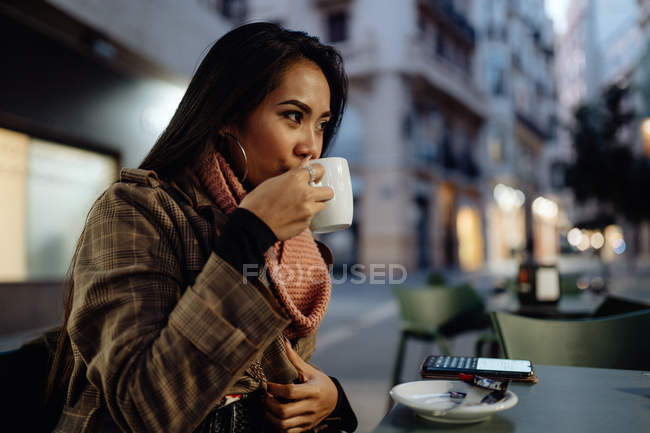 Mulher asiática em roupas da moda saboreando bebida quente fresca e olhando para longe, sentada à mesa e descansando no restaurante de rua à noite — Fotografia de Stock