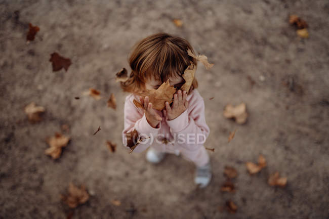 Aktives spielerisches Kind in rosa warmer Kleidung mit geschlossenen Augen vor Vergnügen, das Herbstlaub auf der Wiese im Park wirft — Stockfoto
