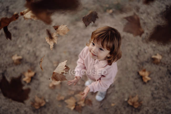 Active playful kid in pink warm clothes throwing up autumn leaves in meadow in park — Stock Photo
