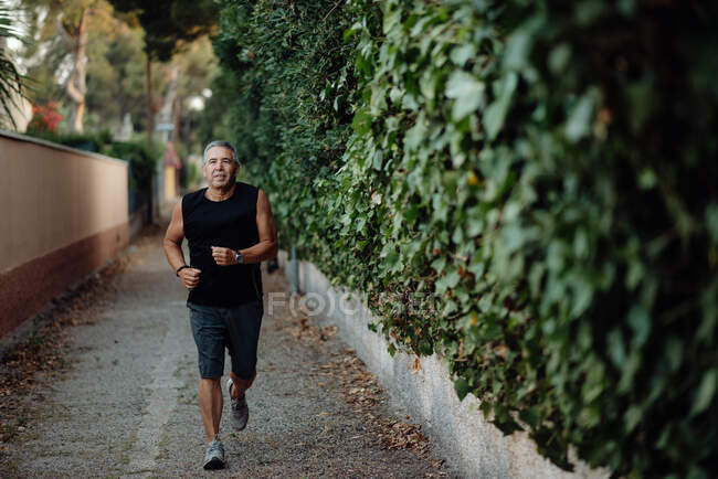 Old man in good shape running on road along plant fence — Stock Photo