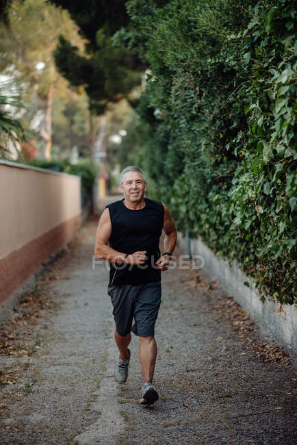 Vieil homme en bonne forme courant sur la route le long de la clôture de la plante — Photo de stock