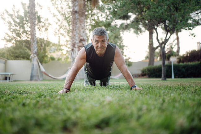 Cheerful elderly man pushing ups on green grass in park — Stock Photo