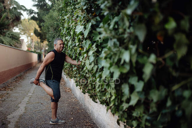 Elderly man stretching leg holding botanical wall in street — Stock Photo