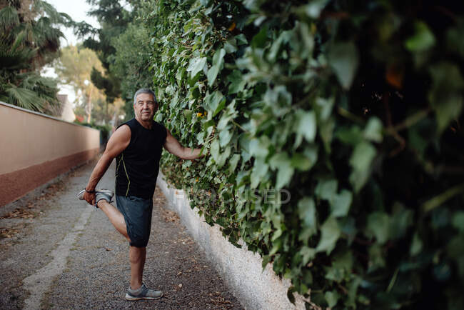 Homem idoso alongamento perna segurando parede botânica na rua — Fotografia de Stock
