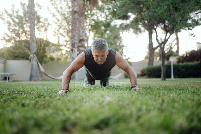 Cheerful elderly man pushing ups on green grass in park — стокове фото
