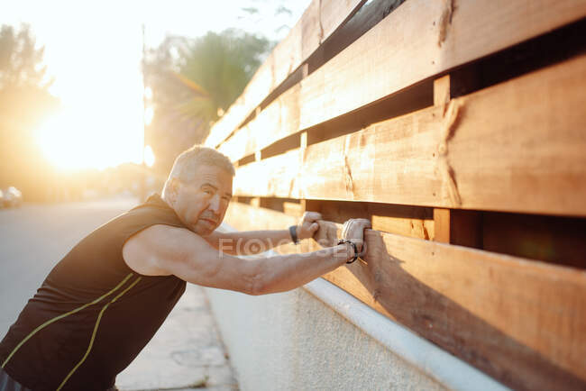 Homme âgé s'étirant sur un mur en bois dans la rue — Photo de stock
