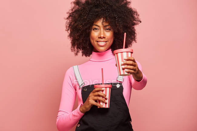 Mulher negra feliz com copo descartável de bebida de pé contra fundo rosa — Fotografia de Stock