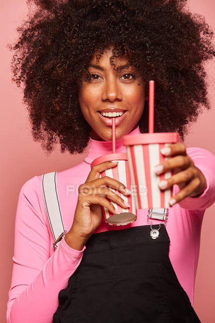 Mulher negra feliz com copo descartável de bebida de pé contra fundo rosa — Fotografia de Stock