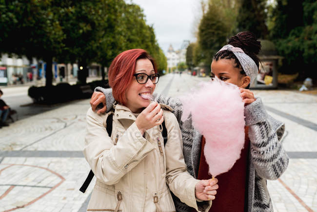 Elegante allegro multietnico ragazza amici a piedi abbracciando e mangiando rosa zucchero filato cotone in strada — Foto stock