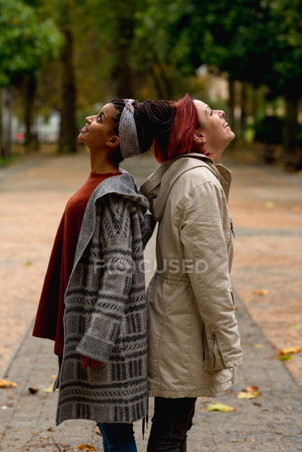 Side view of joyful content stylish multiethnic girl friends enjoying and looking up standing back to back in park — Stock Photo