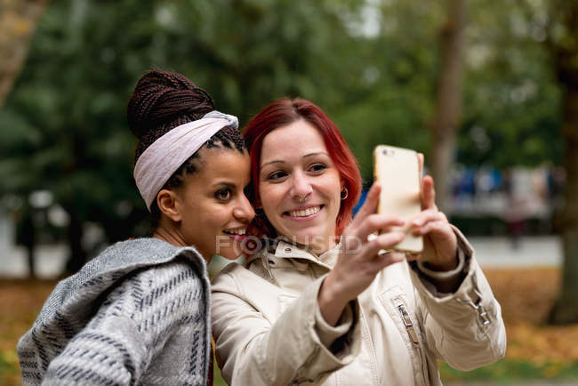 Lovely multiethnic couple of smiling women staying close and taking selfie on mobile phone in autumn park — Stock Photo