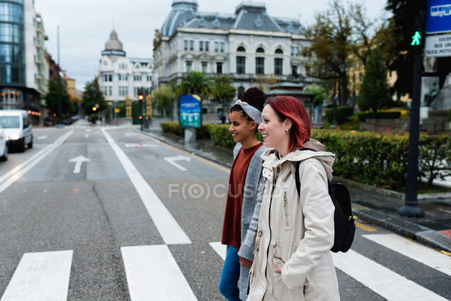 Vista lateral de namoradas multiétnicas elegantes alegres andando e cruzando estrada marcada na cidade em dia cinza — Fotografia de Stock