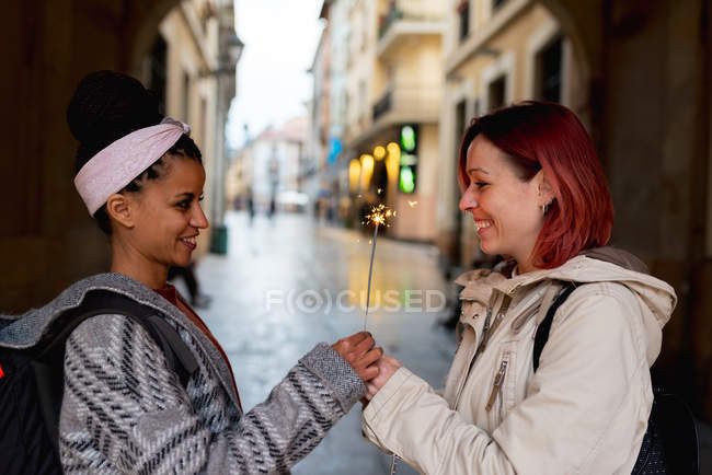 Vista lateral de alegres mujeres multiétnicas alegres permaneciendo juntas y sosteniendo chispa ardiente bajo el arco en la calle - foto de stock