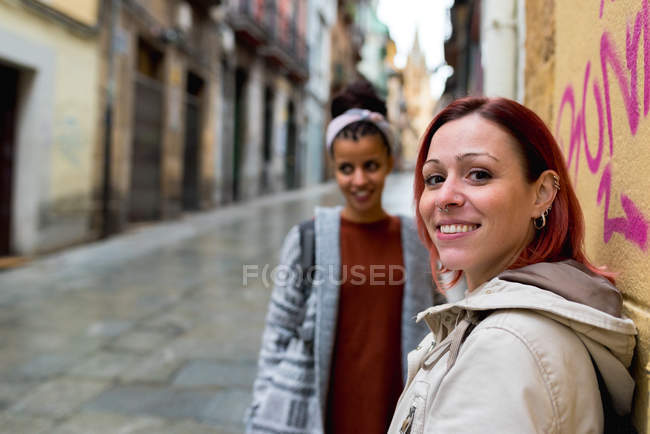 Alegre elegante mujeres multiétnicas caminando y mirando a la cámara cerca de la pared con dibujos en la ciudad - foto de stock
