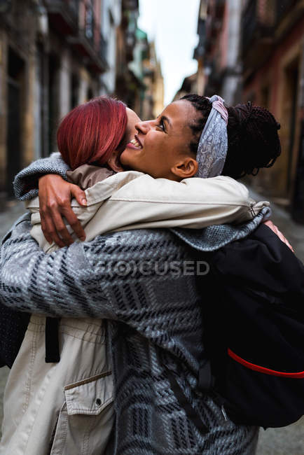 Seitenansicht der schönen multiethnisch lächelnden Frauen, die sich bei einem Treffen in einer engen Straße mit alten Häusern umarmen — Stockfoto
