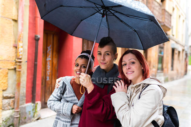 Zufriedene multiethnische Frauen, die unter einem großen schwarzen Regenschirm spazieren und vor dem Hintergrund roter alter schäbiger Wände in die Kamera schauen — Stockfoto