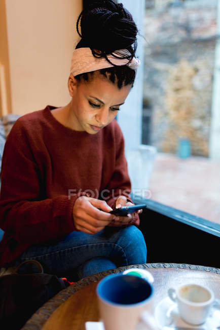Mulher afro-americana elegante pensativo surfar telefone móvel sentado com pernas cruzadas à mesa com café no café — Fotografia de Stock
