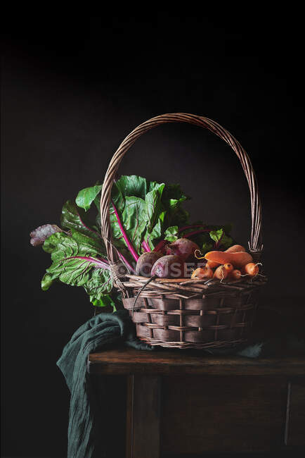 Natura morta di cesto di vimini con manico pieno di verdure fresche da giardino su tessuto e tavolo di legno su sfondo nero — Foto stock