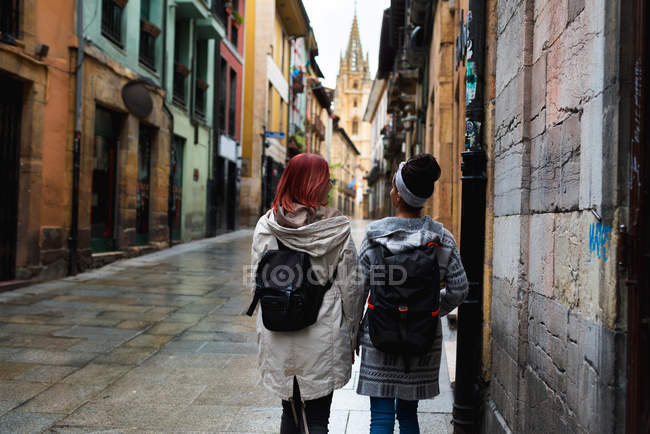 Vista trasera de mujeres turísticas activas con mochila caminando por calles vacías en la ciudad - foto de stock