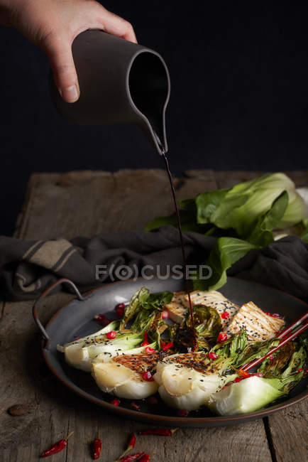 Person verschüttet Sojasauce aus Krug auf köstlichem Bok Choy Salat mit Fisch auf Holztisch — Stockfoto