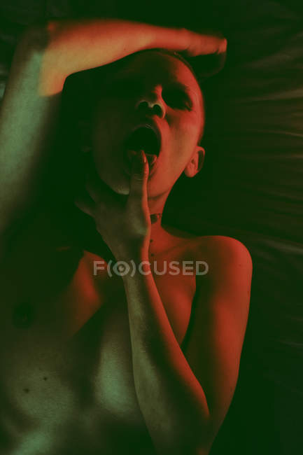 Sexual quente apaixonado mulher nua com a cabeça raspada deitada na cama com o dedo na boca em estúdio escuro com luz vermelha — Fotografia de Stock