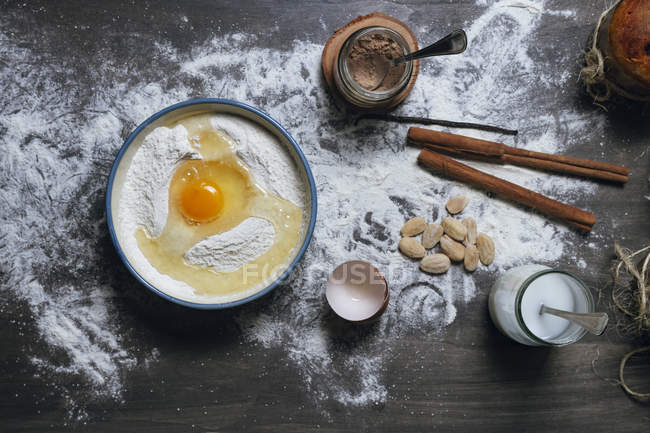 Vista superior de los ingredientes para la receta de panettone que incluye un tazón con harina y huevo y frascos con cacao en polvo y leche colocada en la mesa espolvoreada con almendras y especias - foto de stock