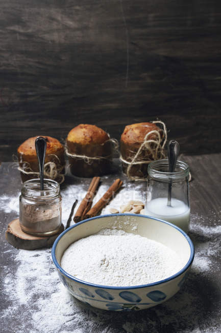 Bowl with white flour and glass jars with cocoa powder and milk placed on table next to spices and baked panettones — Stock Photo